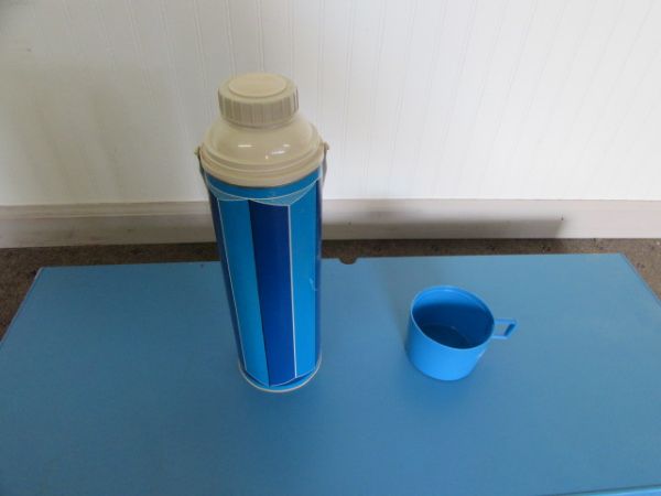 BLUE FOLDING CAMP TABLE WITH ATTACHED BENCHES & A BLUE THERMOS.